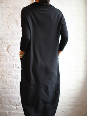 3/4 back view of woman modeling simulacra's black funnel neckline cocoon dress with pockets